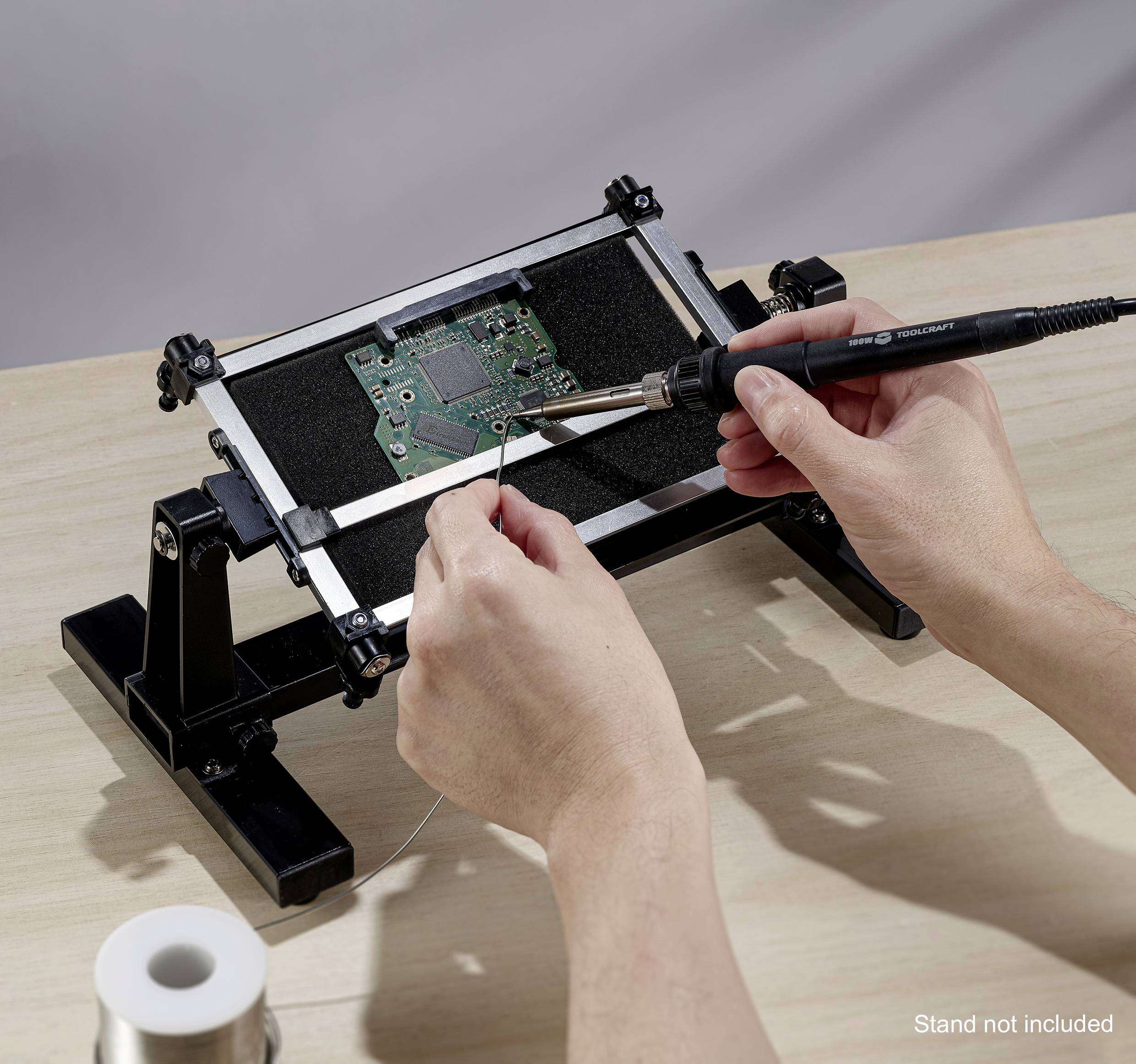 A person is soldering a circuit board with a soldering iron on a table. One hand is holding the soldering iron, the other is guiding wire to the solder point.