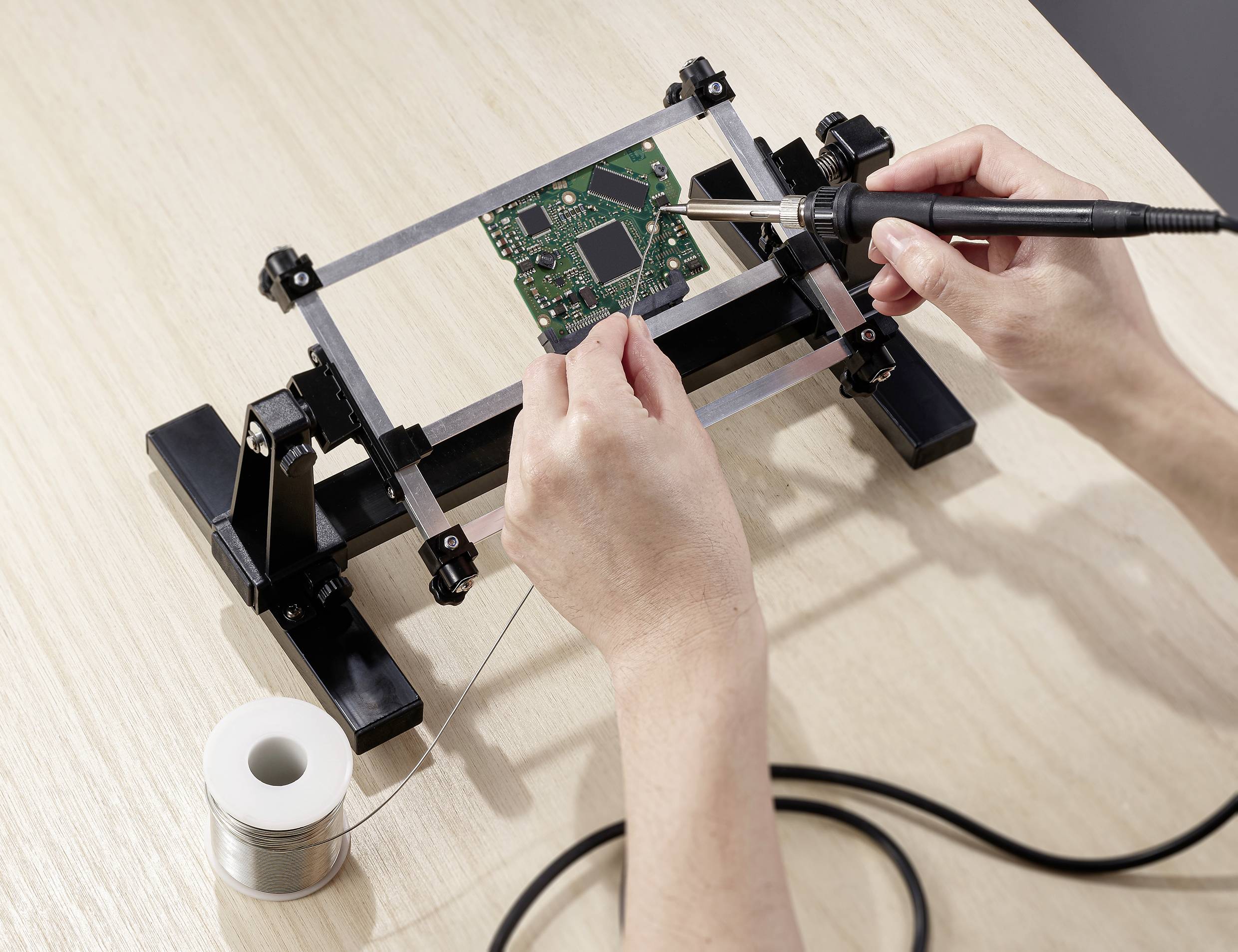 A person is soldering a green circuit board with a soldering iron, held in a stand, on a wooden table.