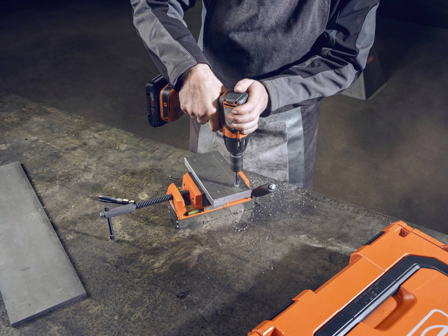 A person in work attire is using an orange drill to machine a piece of metal in a clamp on a workbench.