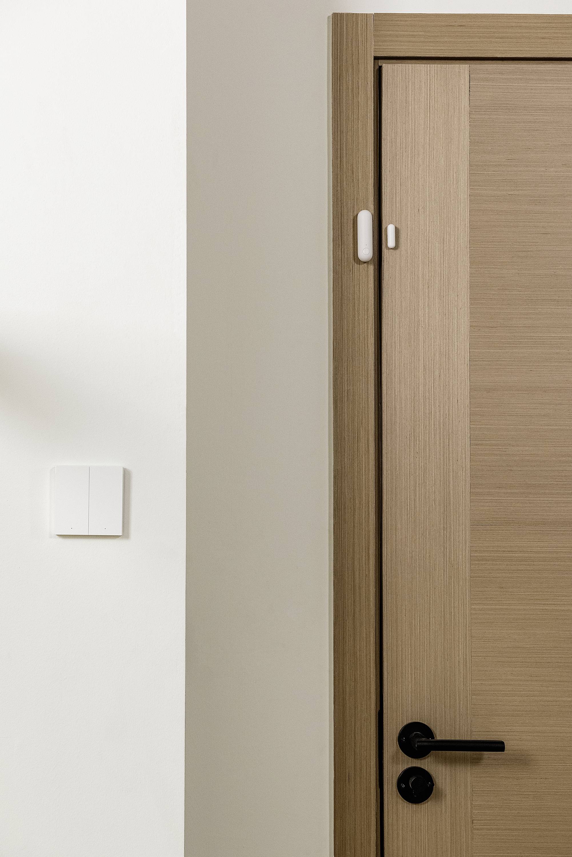 A half-open wooden door with a black door handle next to a white wall, featuring a light switch and a sensor on the door frame.