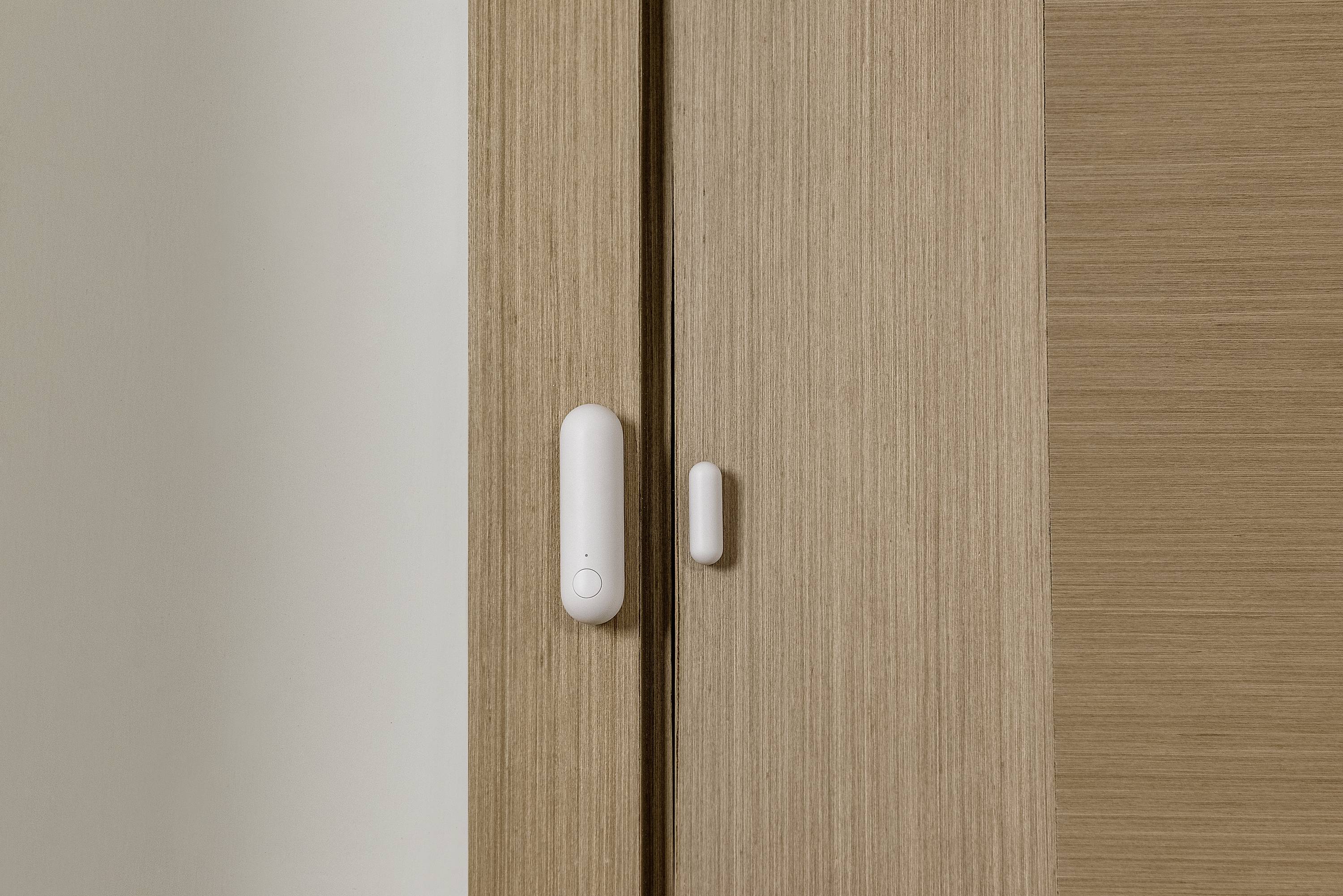 A window or door sensor in white is attached to a wooden door frame to detect movement or opening.