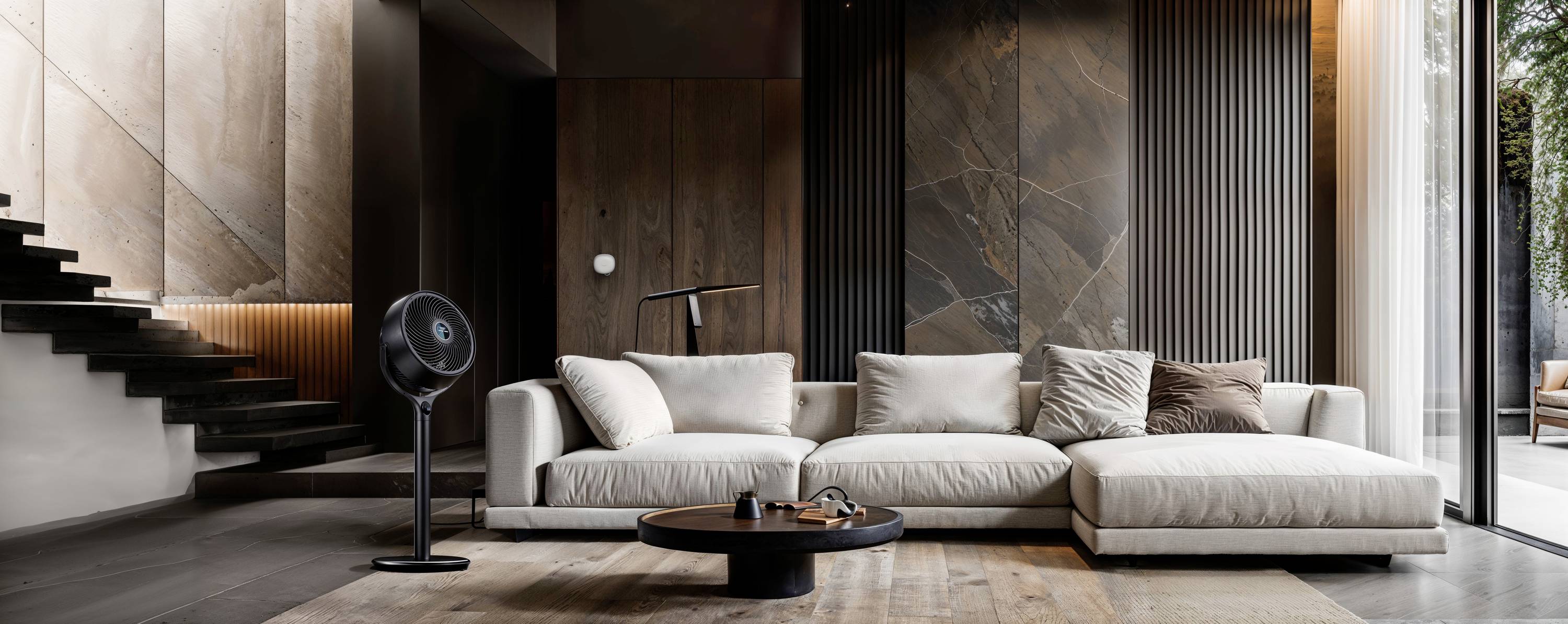 Modern living room with a beige sofa, round coffee table and floor lamp. Wooden walls and staircase on the left side, large windows on the right.