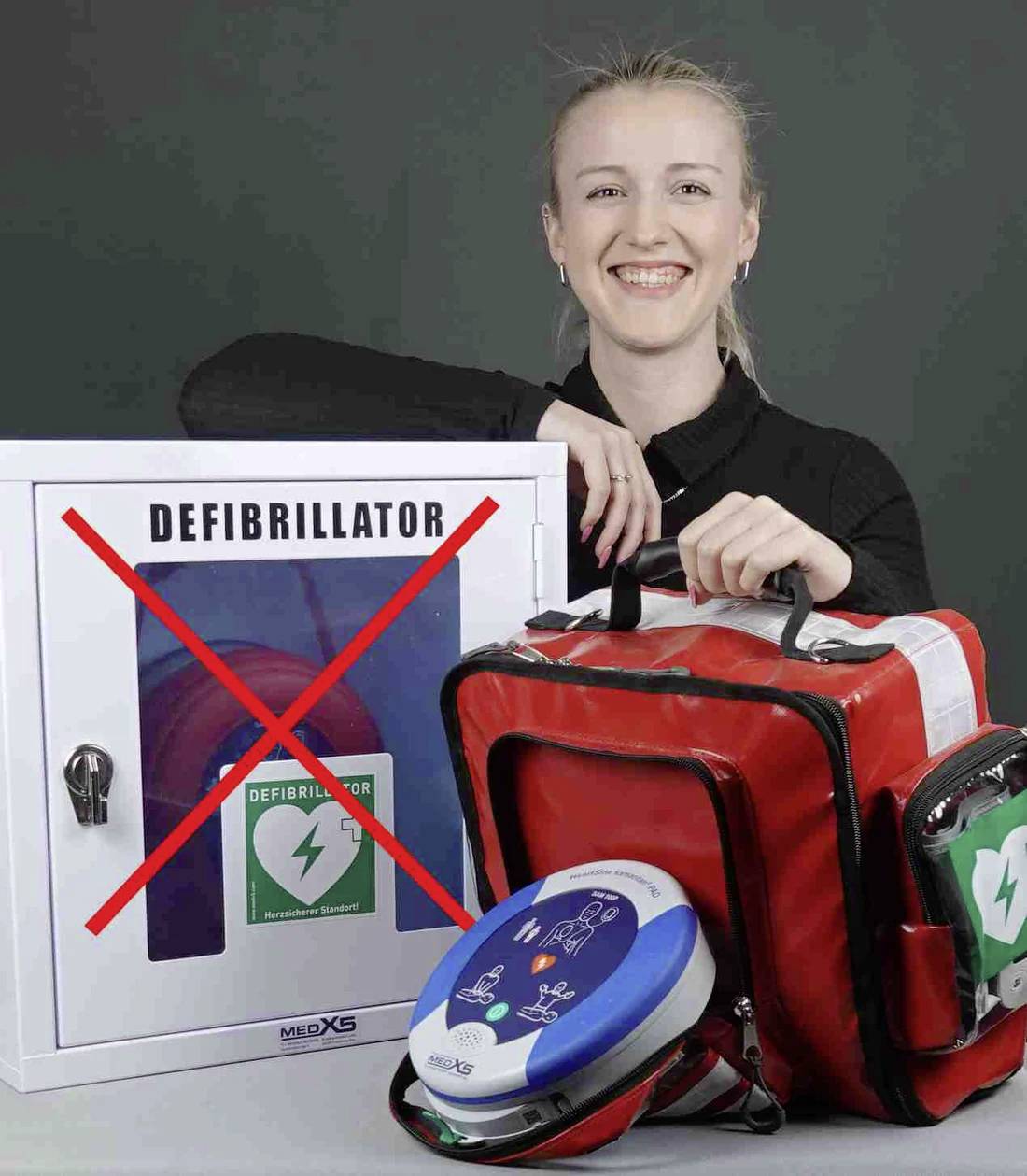 A person is smiling next to a defibrillator cabinet marked with a red X, holding a red emergency bag with a portable defibrillator.