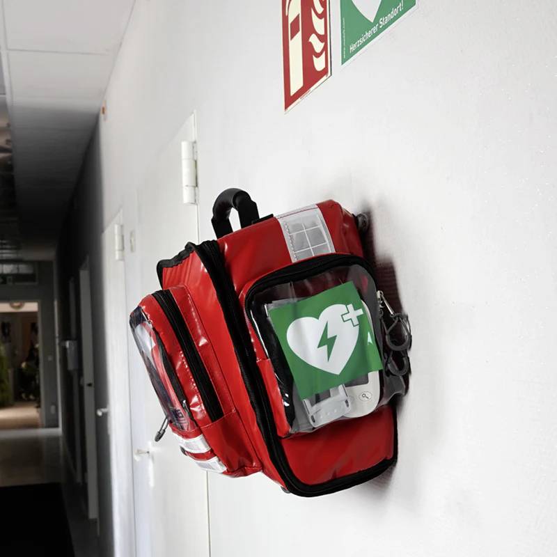 A first aid backpack with an AED symbol is hanging on the wall beneath a fire extinguisher sign in a bright corridor.