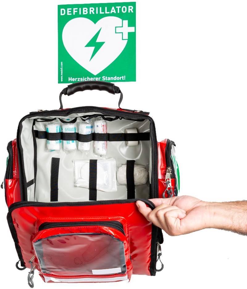 A red emergency rucksack with first aid equipment is opened. A sign indicates 'Defibrillator' at a safe location.