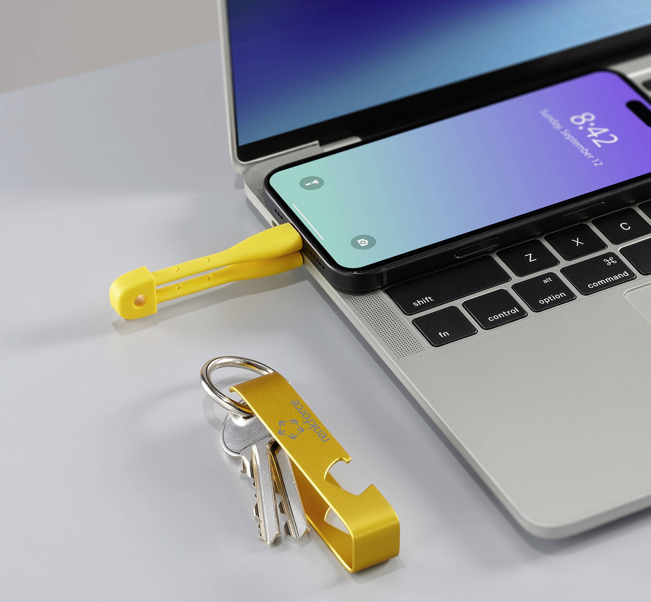 An open laptop with a smartphone resting on the keyboard, a USB stick plugged in, and a yellow keyring with keys beside it on the table.