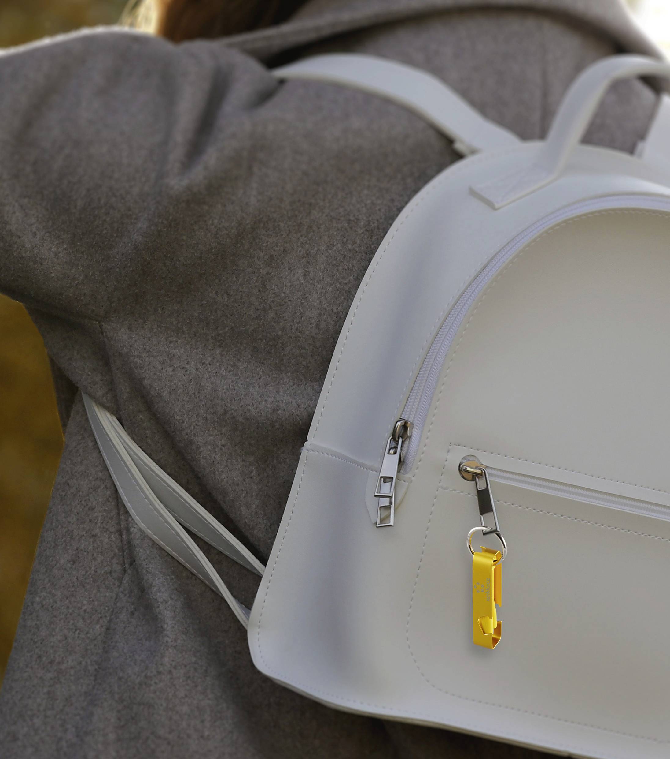 A person in a grey coat is wearing a white rucksack; a yellow keyring is attached to it.