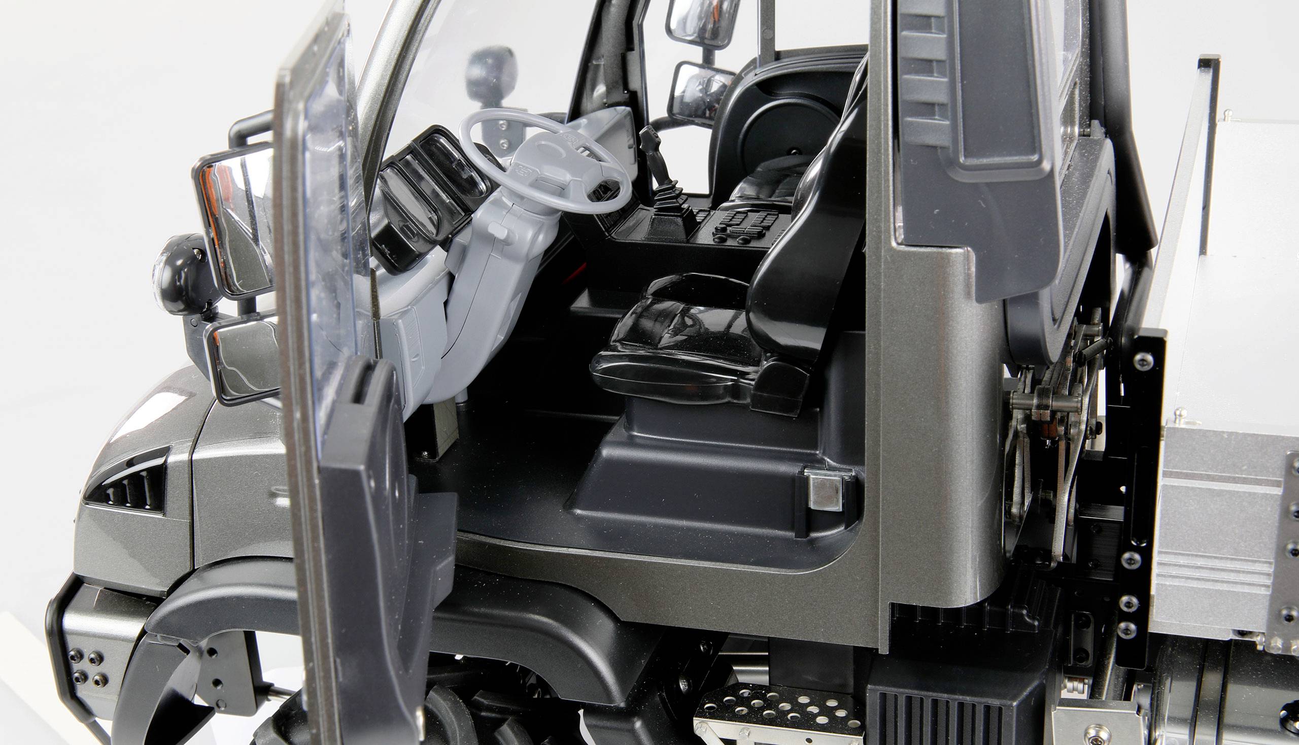 Interior view of a vehicle: open cab with steering wheel, two seats, dashboard and open door, in black and grey colour scheme.