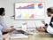 A group of people in a meeting are examining diagrams of product sales data on a screen and laptops.