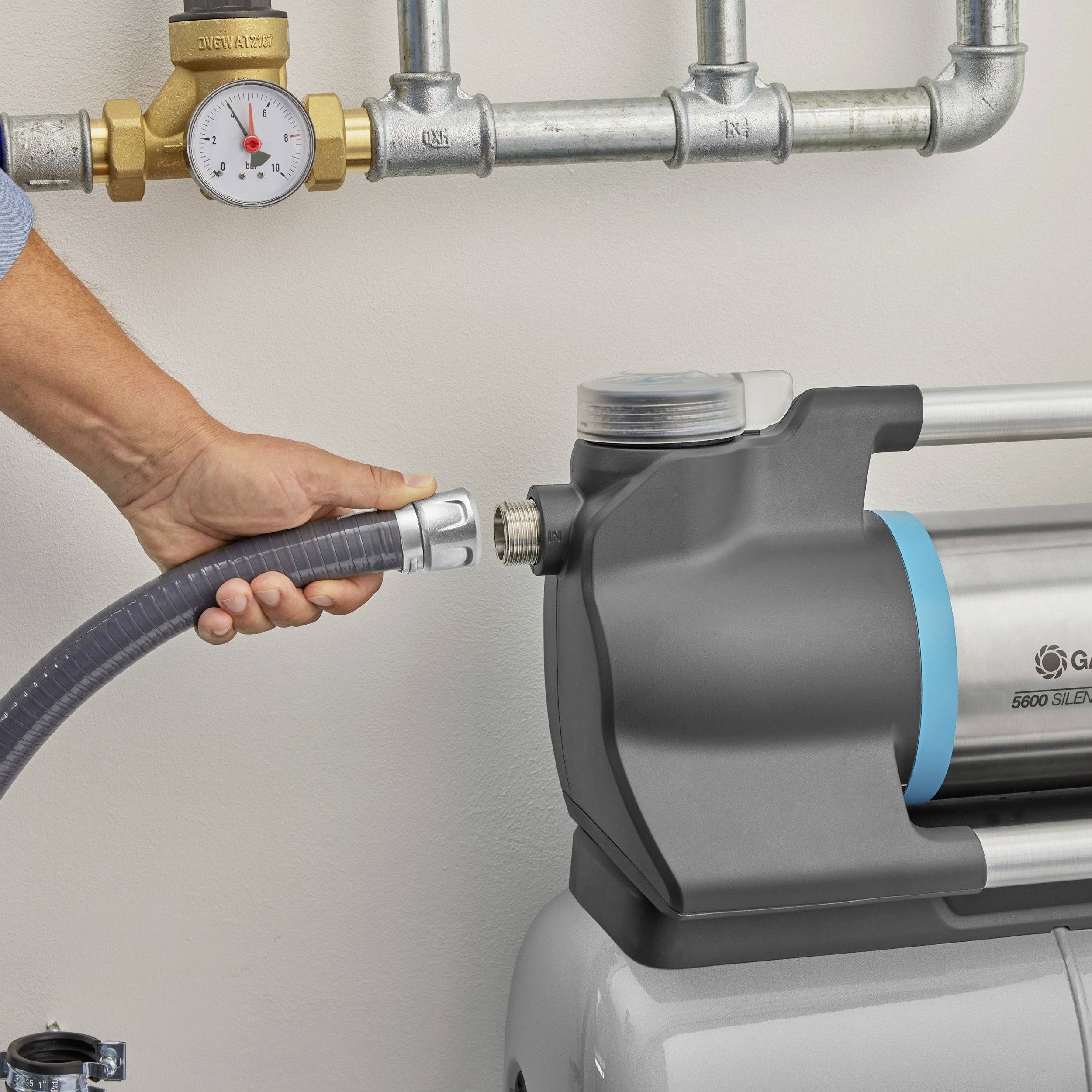 A person is holding a black hose connected to a metallic pump. Pipes and a pressure gauge are visible in the background.