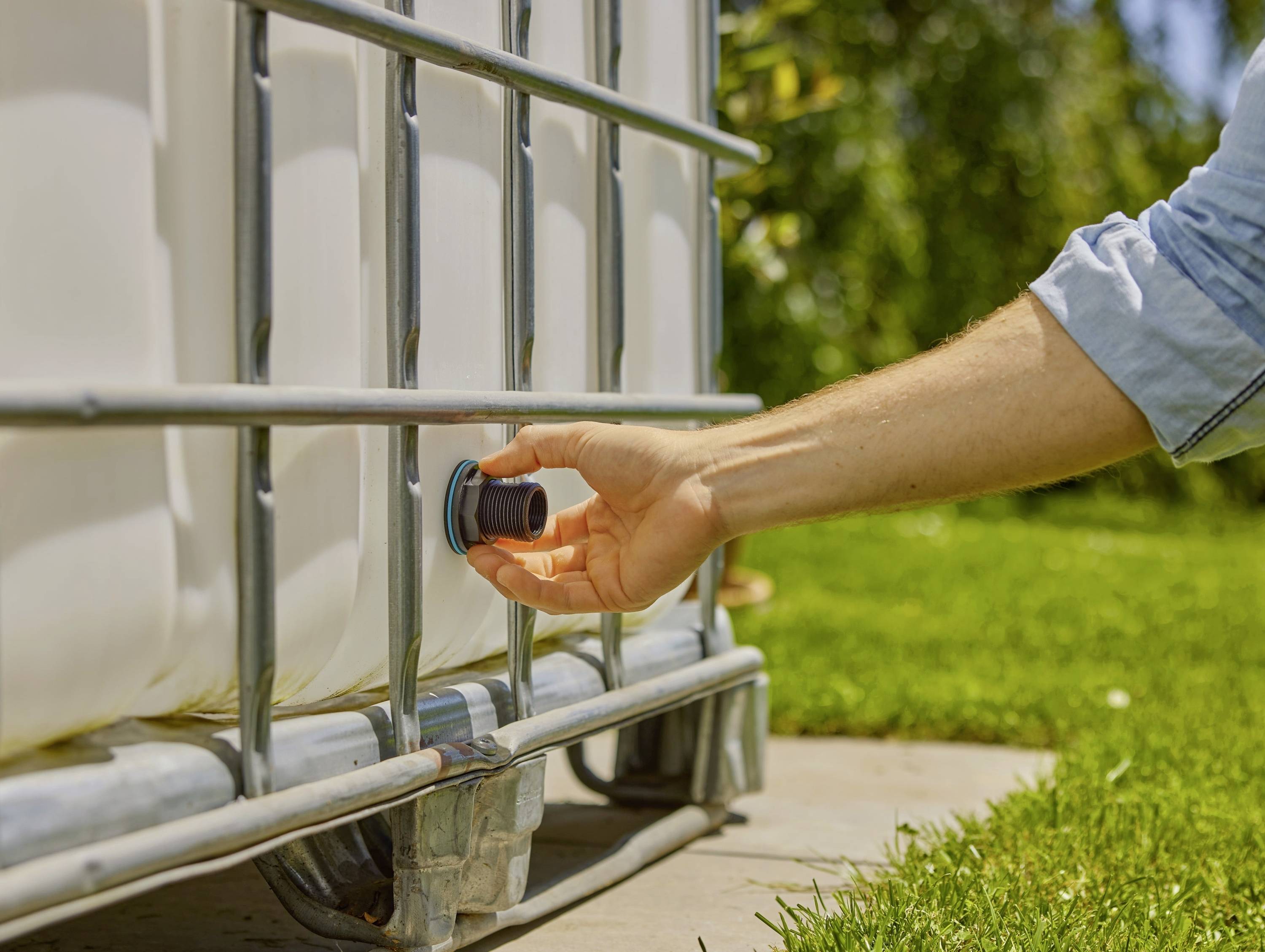 A hand turns the outlet tap of a large white water container outdoors. Green vegetation is visible in the background.