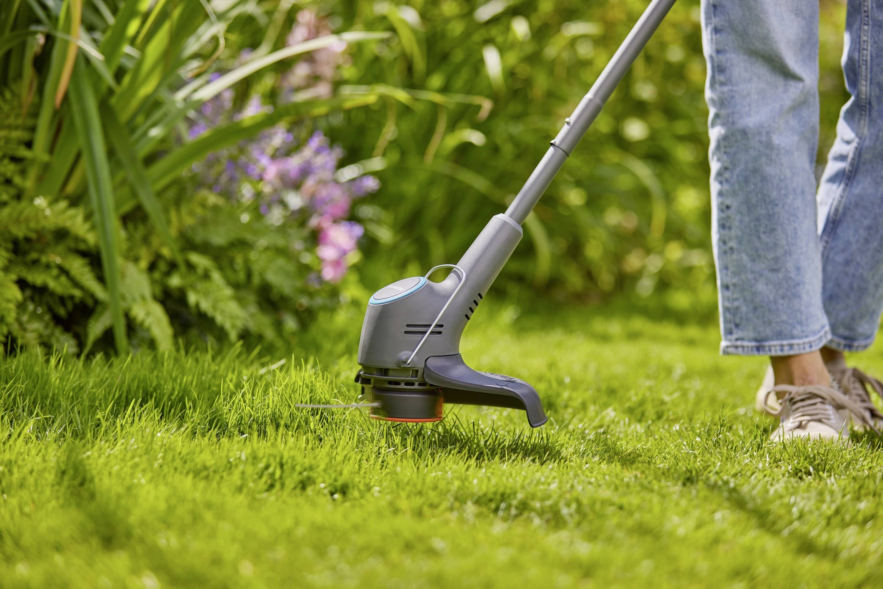 A person is trimming the lawn with an electric lawn trimmer in a garden. Flowers and green plants are visible in the background.