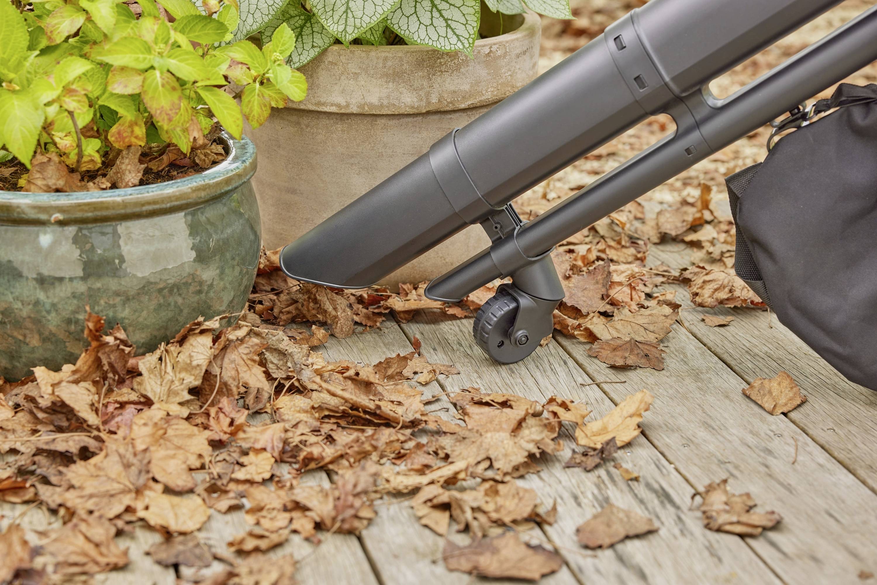 A leaf blower removes dry leaves from a wooden decking. Potted plants are positioned beside the foliage.
