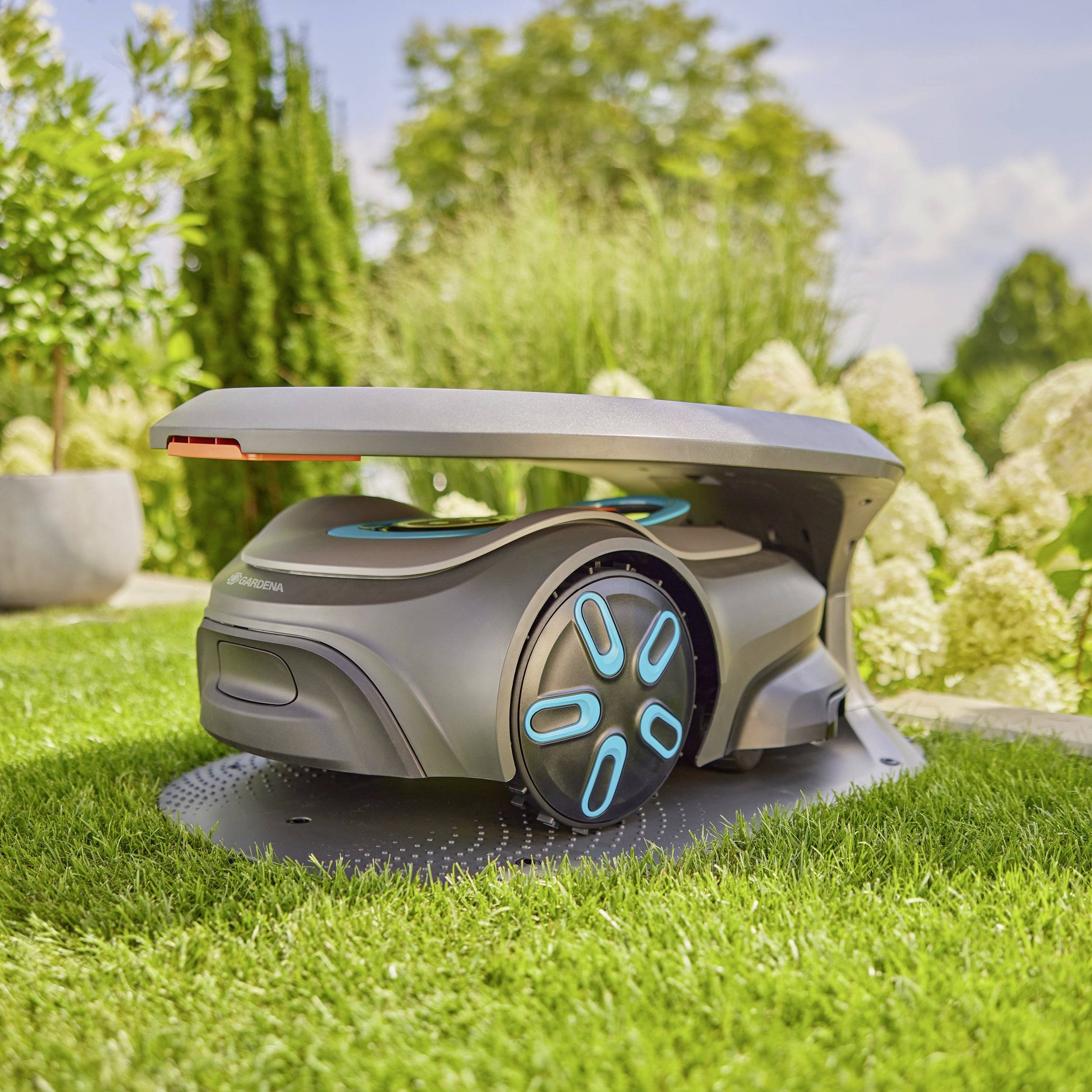 An automatic lawnmower on a lawn area on a sunny day. Bushes and flowers are visible in the background.