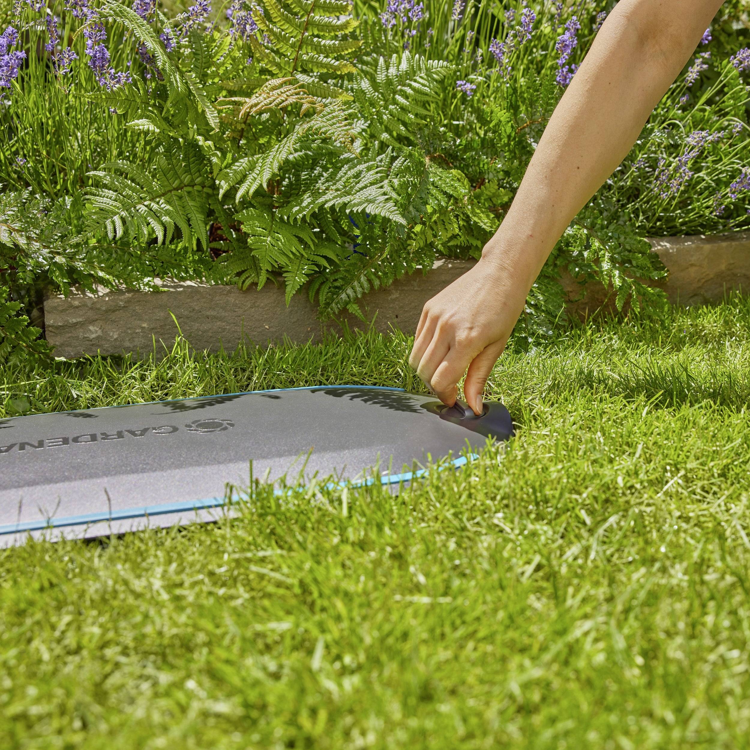 A person touches a blue GARDENA ground mat in the garden, surrounded by green plants and purple flowers, on a sunny day.
