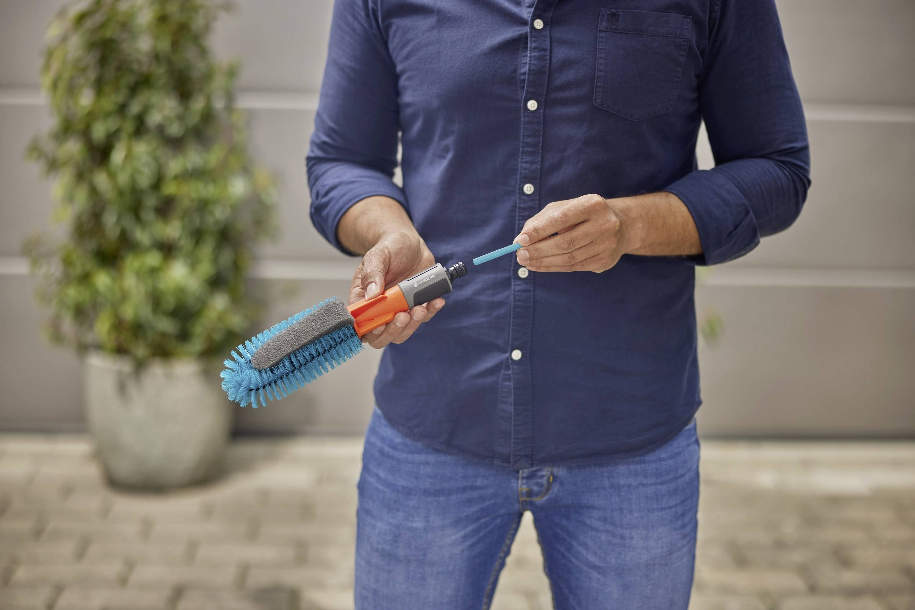 A person wearing blue clothing stands outdoors holding a door handle brush. Plants and a wall are visible in the background.
