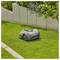 A robotic lawn mower autonomously trims a small section of grass in a garden, bordered by concrete planters and various plants.