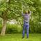 A man is standing on the lawn and trimming branches from a tree with a pair of secateurs. Surrounded by green foliage and dense vegetation.