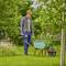 A man in the garden is pushing a wheelbarrow with branches. He is wearing casual clothing and holding garden shears, surrounded by trees.