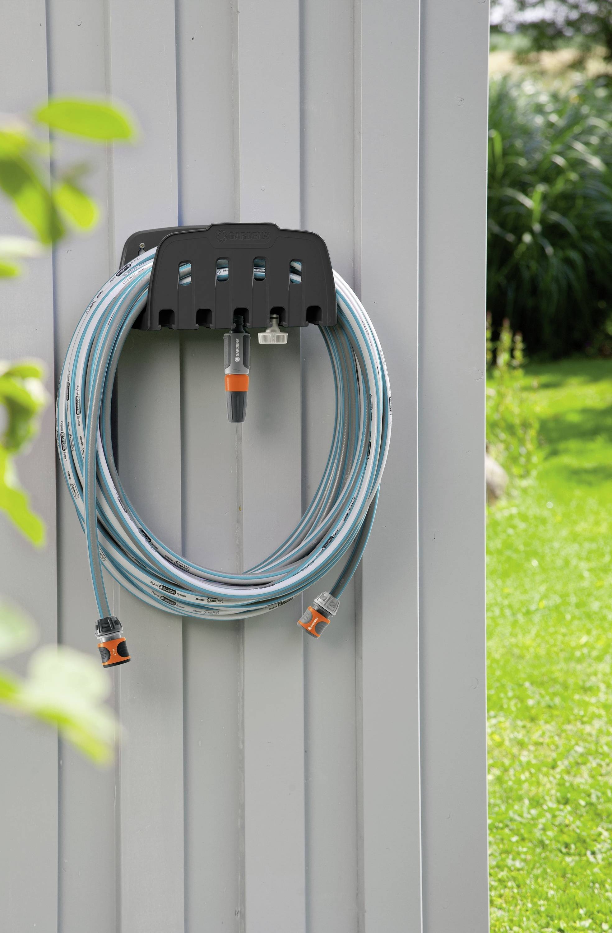 Garden hose, neatly hung on a mounting bracket on a grey wall, with green lawn and plants in the background.