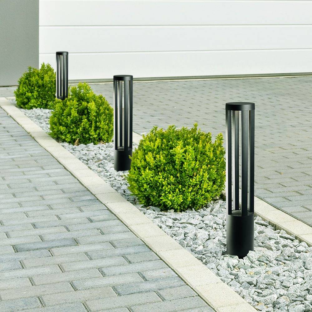 Three black outdoor lights along a paved path, flanked by green bushes and pebbles, in front of a white wall.