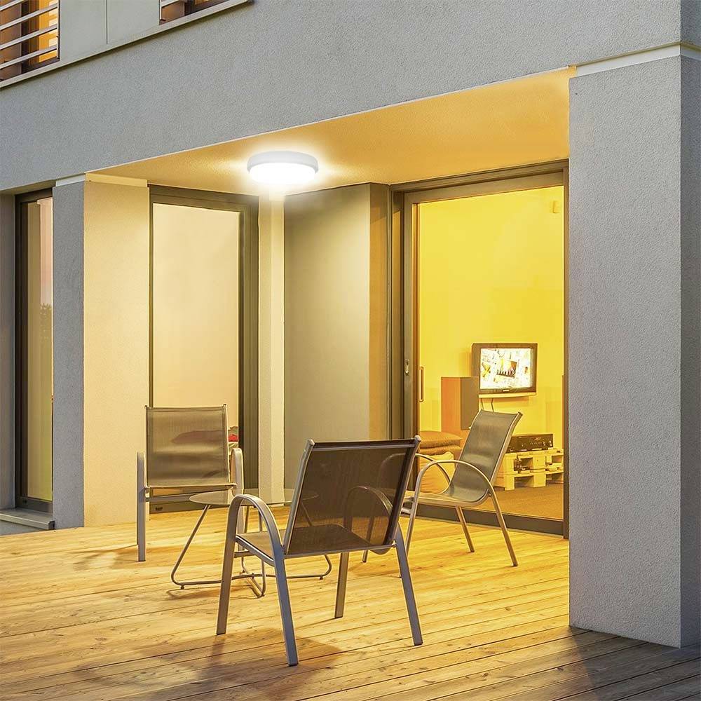 Balcony of a modern house at night, illuminated by an outdoor lamp; two chairs and a table in the foreground, window revealing a lit interior.