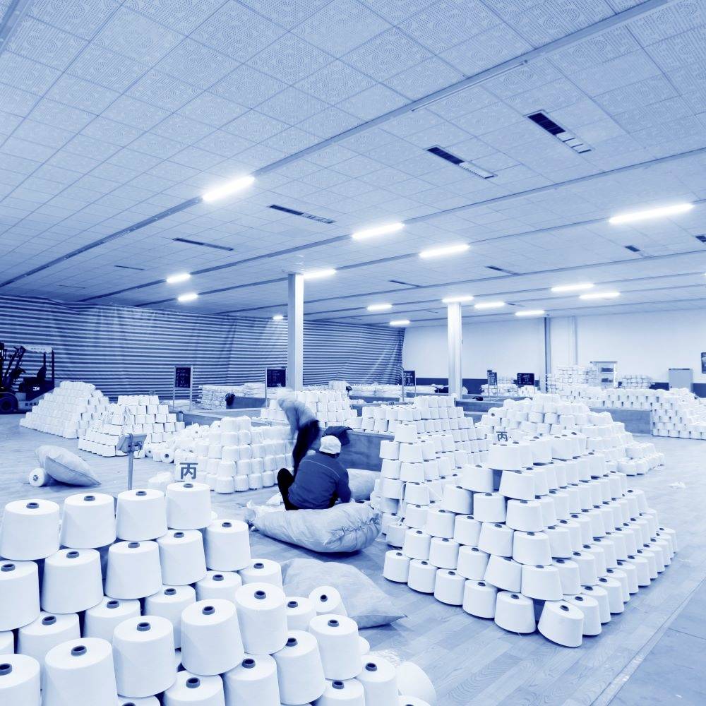 A large storage area with stacked rolls, presumably paper or yarn. Two people are working between the stacks. The room is brightly lit.