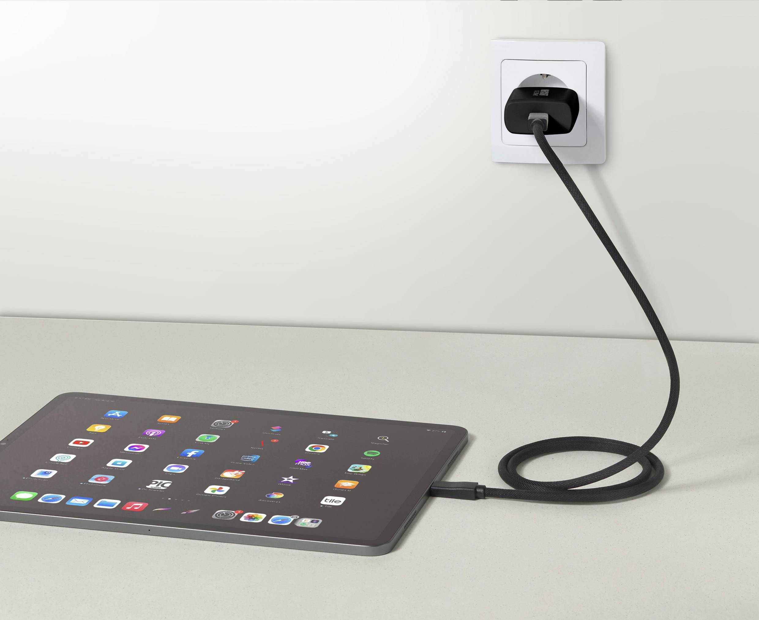 A tablet is lying on a table and is connected to a socket with a cable. It displays numerous colourful app icons on the screen.