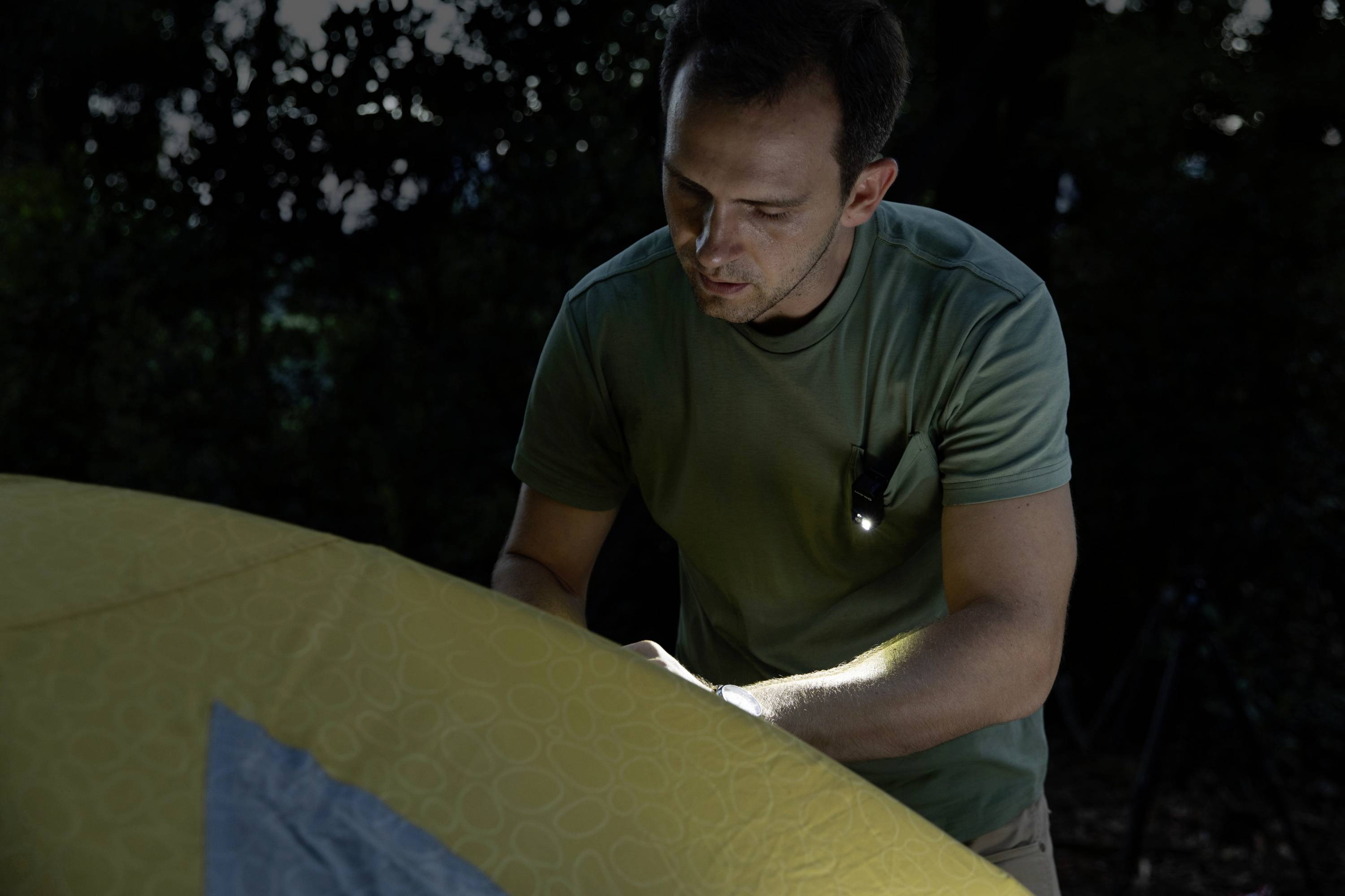 A man is pitching a tent outdoors in low light. He is wearing a green T-shirt and using a torch for illumination.