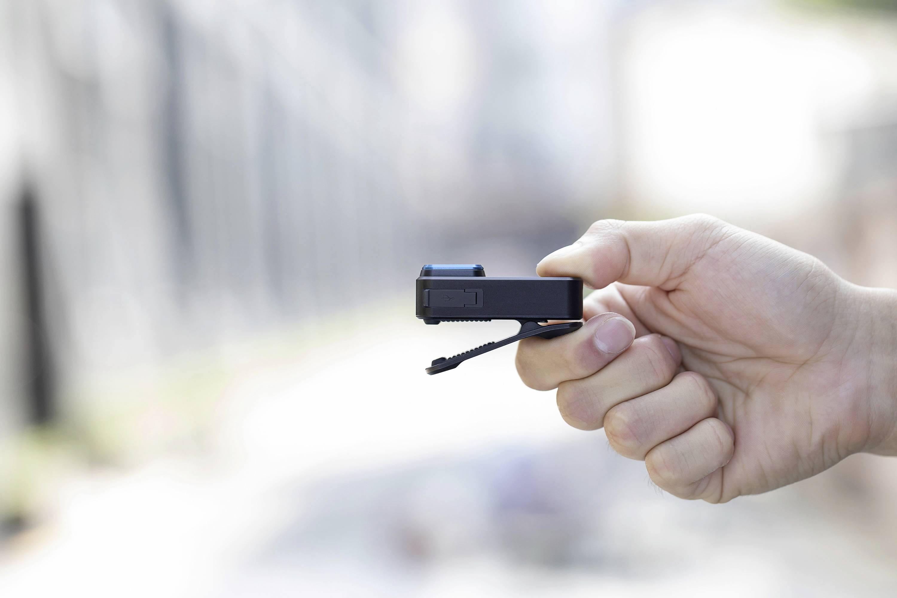 A hand is holding a small black camera with a strap outdoors. The background is blurred.