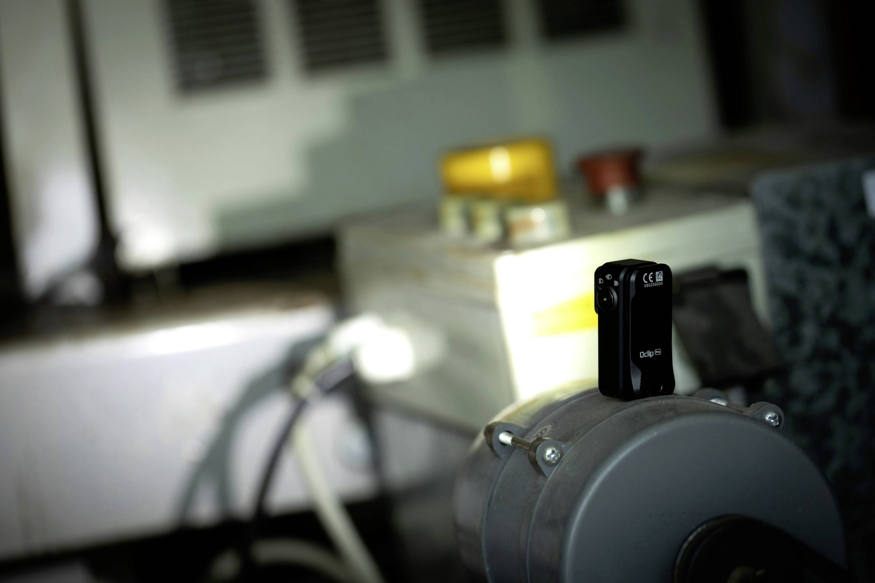 A small black camera is mounted on a machine, surrounded by a grey background with yellow and red buttons.