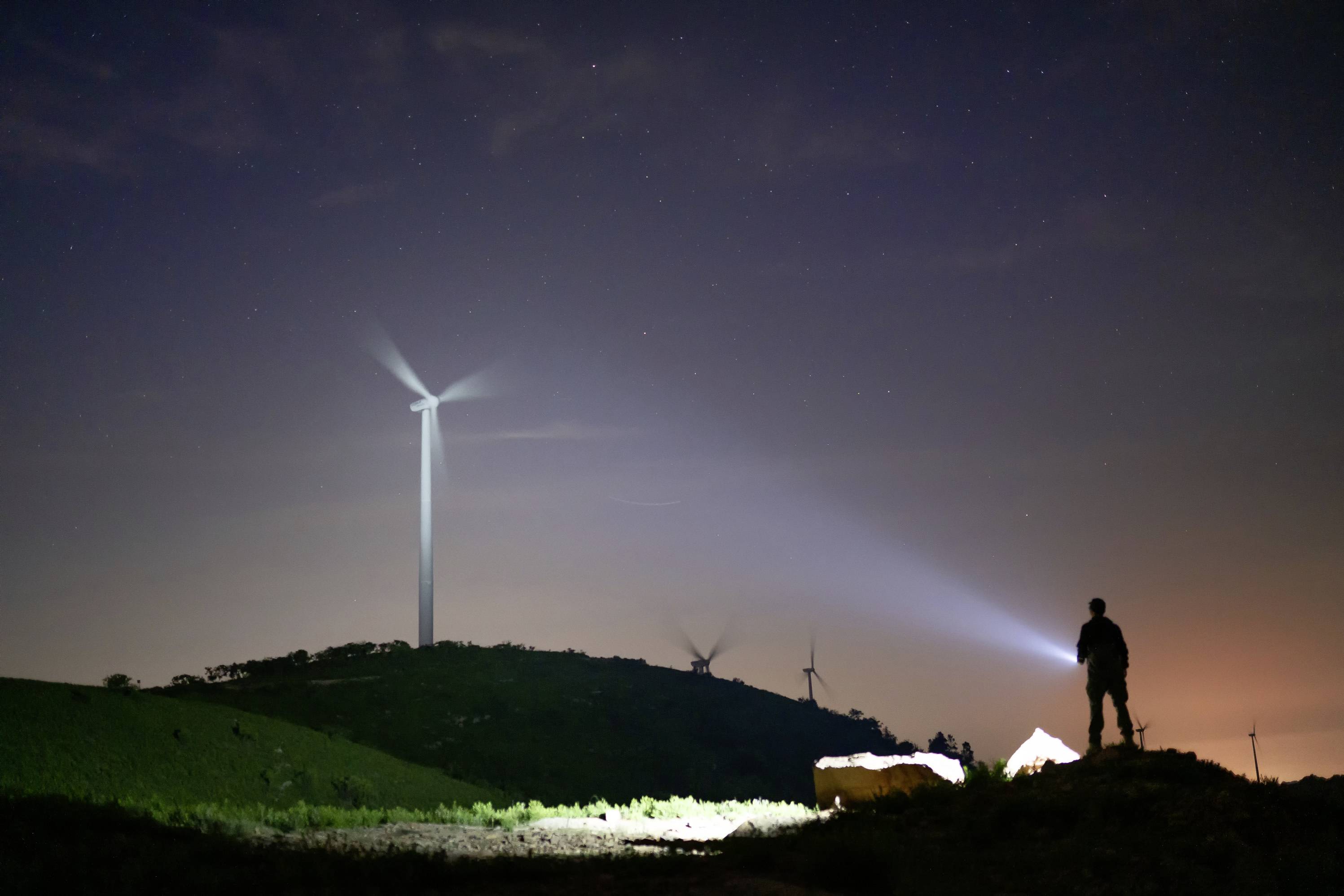 A person stands on a hill at night with a torch, illuminating an area. Wind turbines are operating in the background.