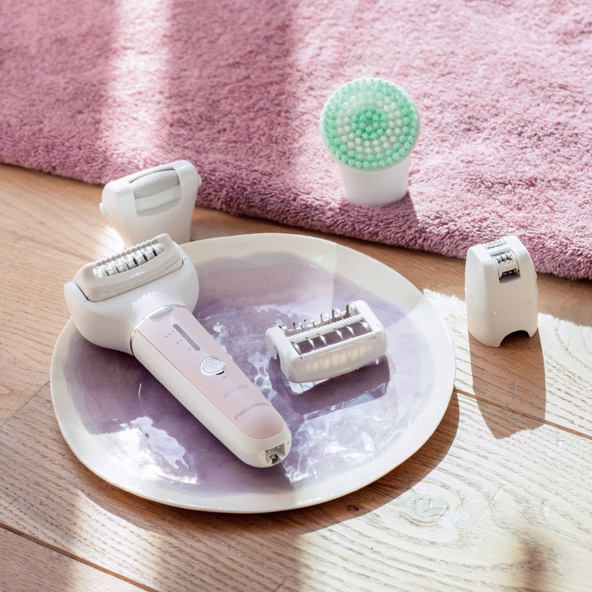 Electronic epilator with multiple attachments rests on a round plate on a wooden floor; pink towel in the background.