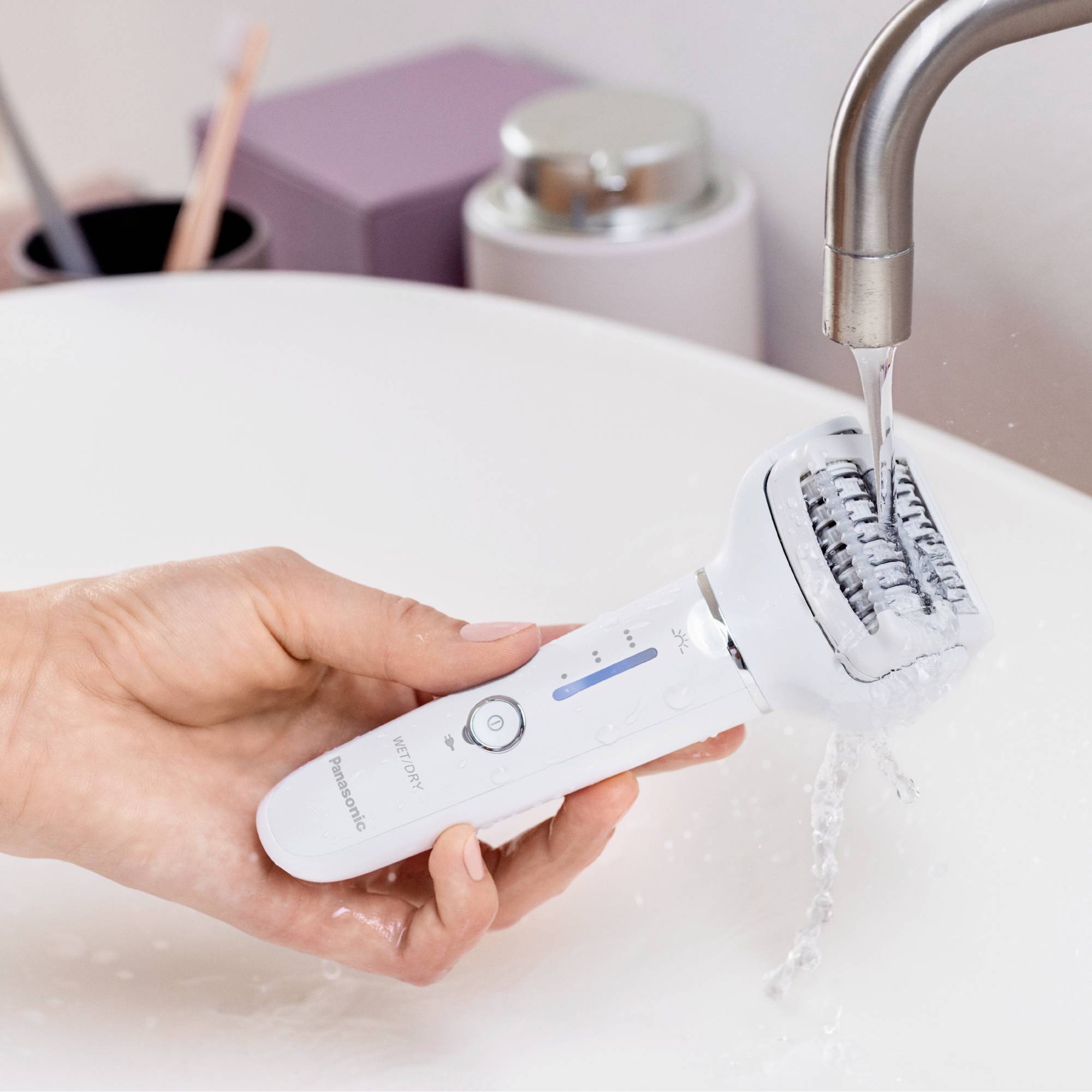 A hand holds an electric epilator under running water in a sink for cleaning.