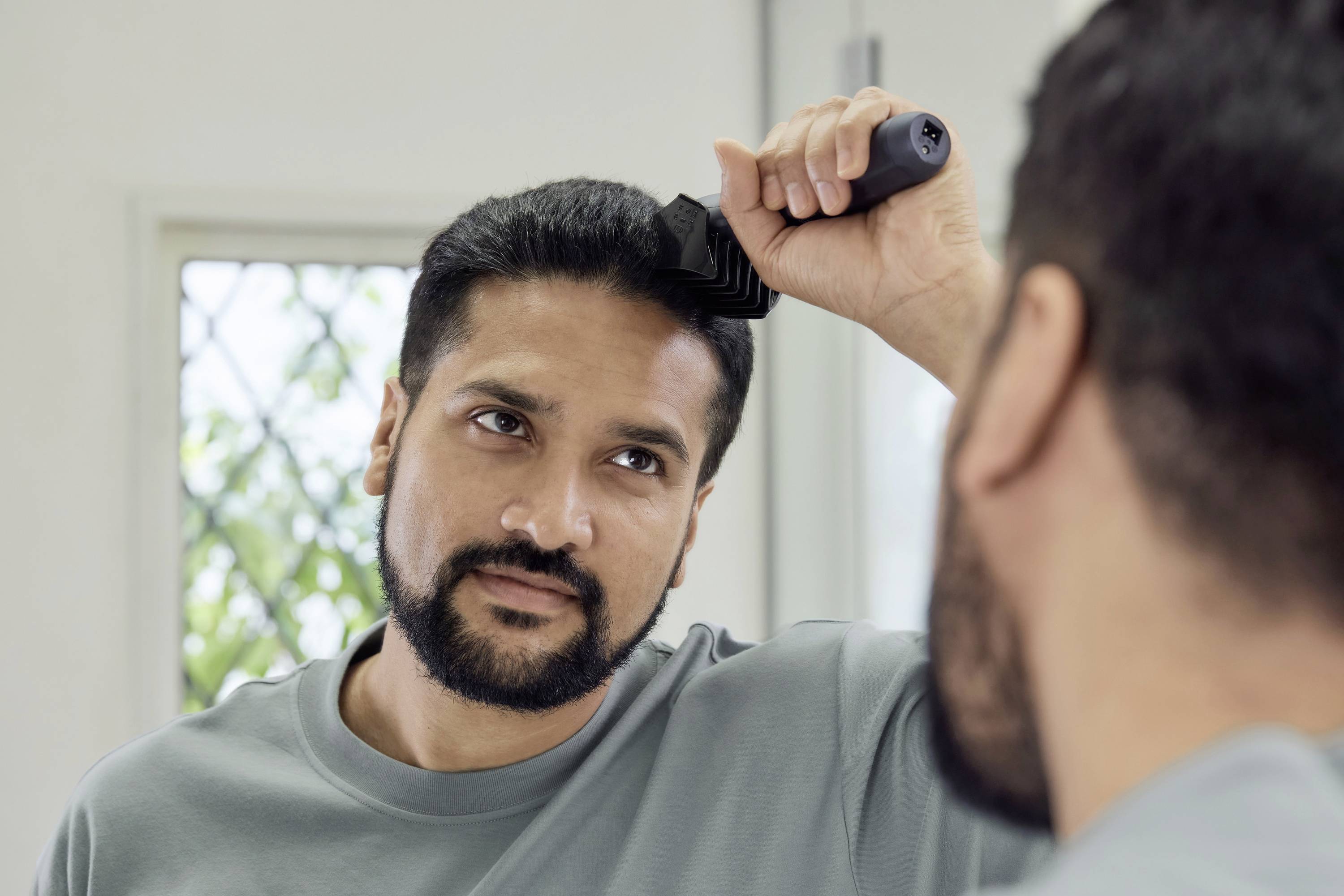 A man is combing his hair in front of a mirror. He is wearing a grey shirt. In the background, there is a window with a slightly blurred view outside.