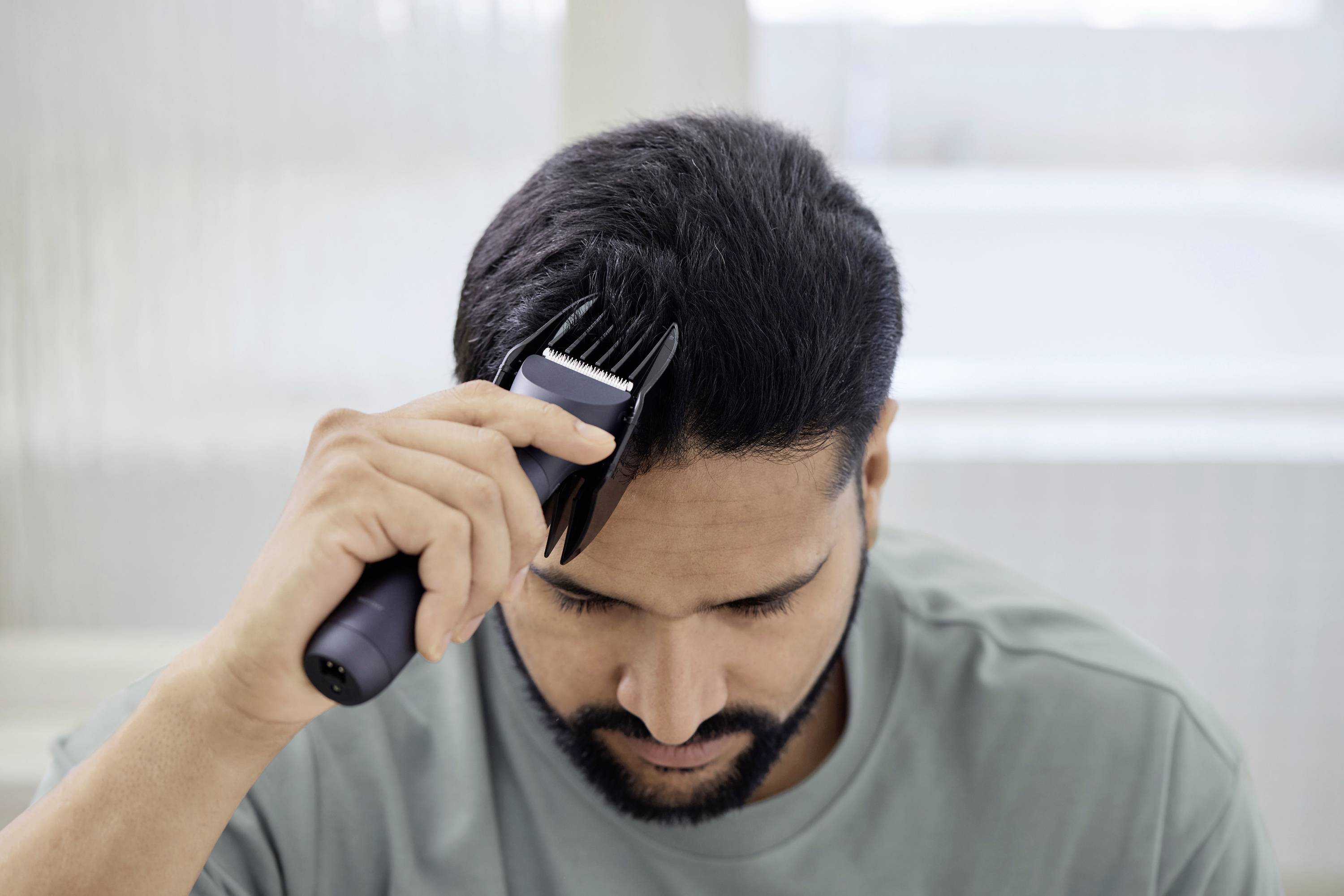 A man is cutting his hair with an electric hair clipper. He is focused on his task in the bathroom.