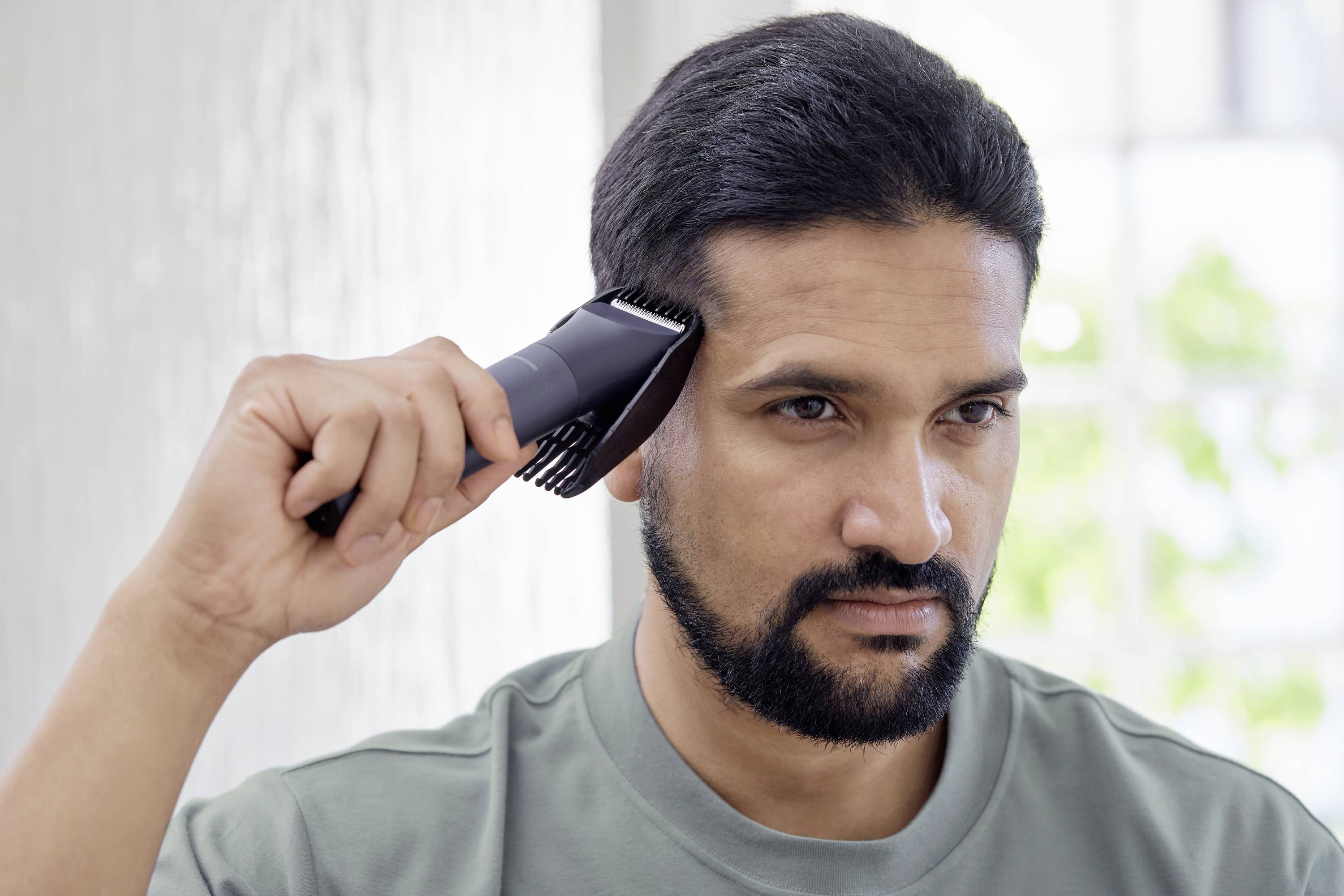 A man with a beard combs his hair with an electric hair trimmer in front of a light background.