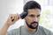 A man with a beard combs his hair with an electric hair trimmer in front of a light background.