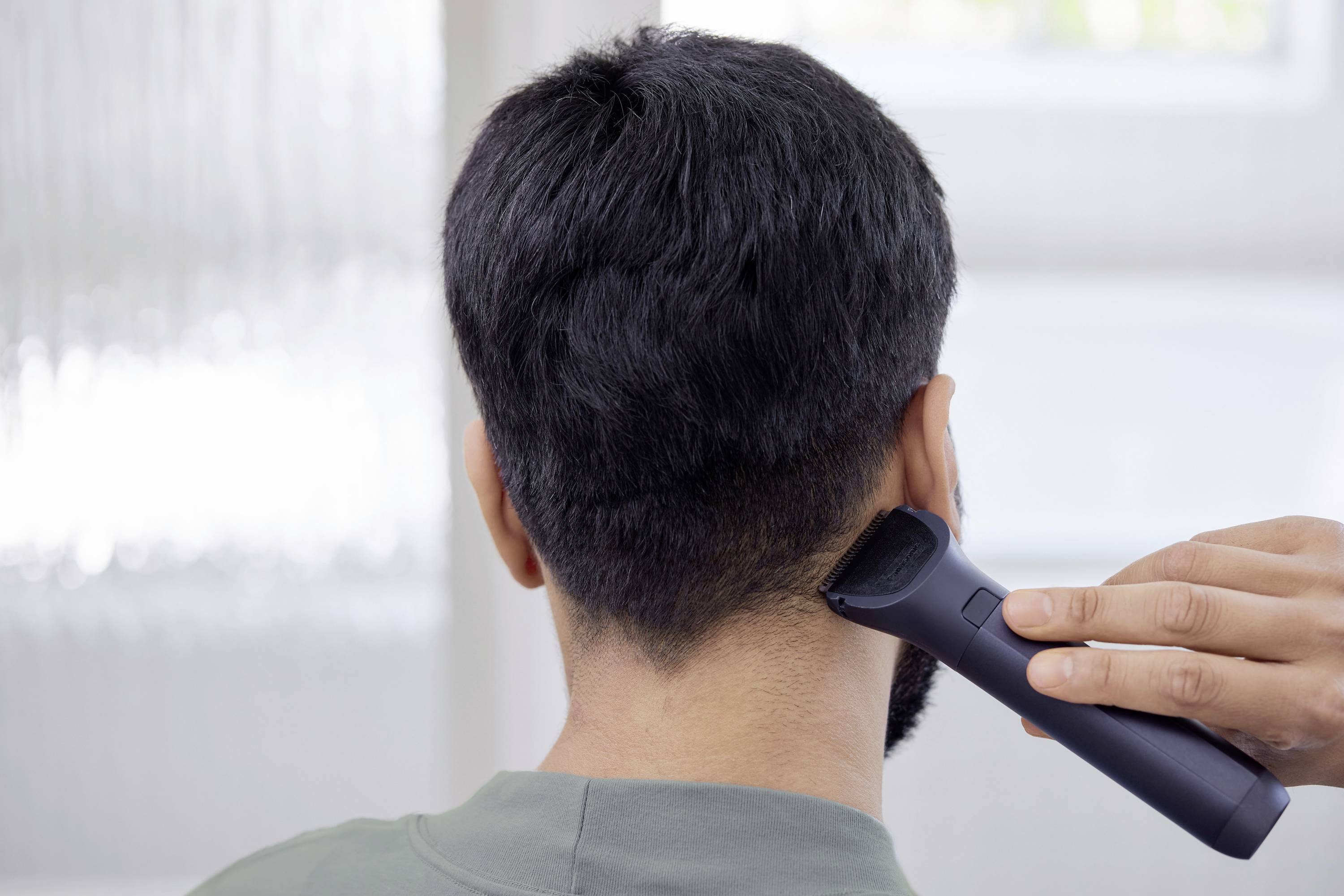 A person is shown from behind while their hair is being trimmed with an electric razor. Focus is on the nape of the neck.