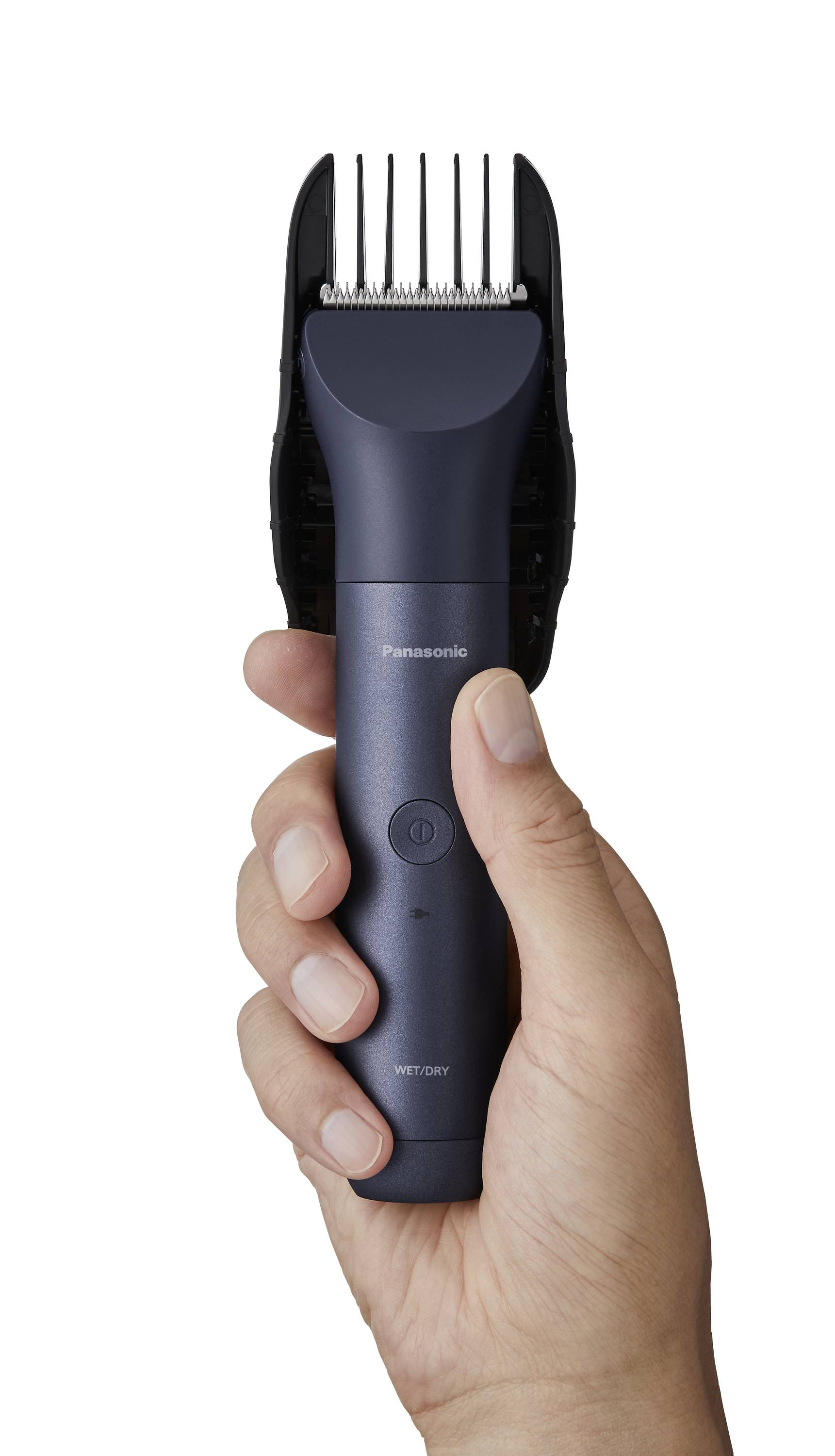 An electric hair clipper is held in the hand. The device is dark grey with silver blades and has an ergonomic shape.