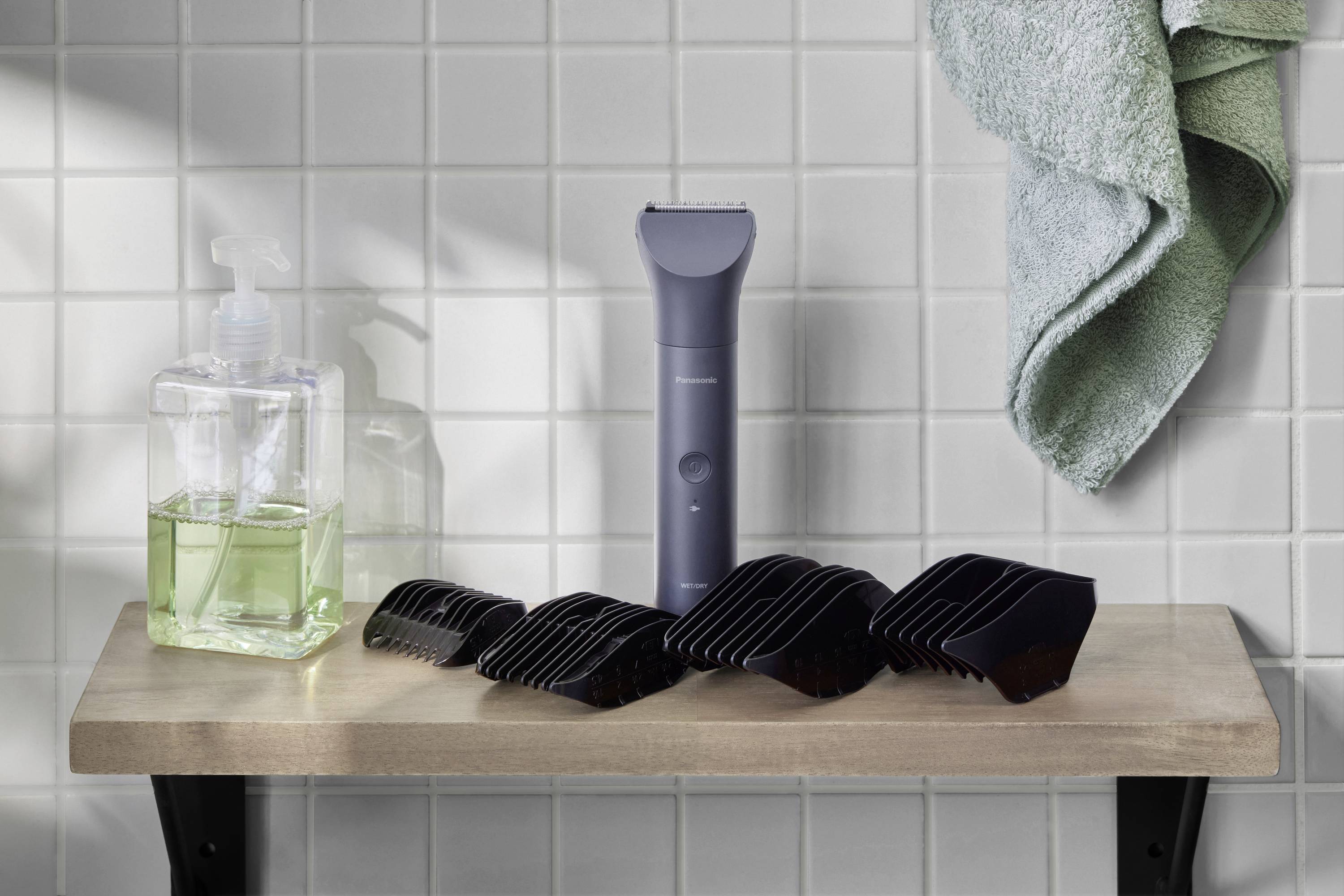 Electric razor and black attachments on a shelf in the bathroom, next to a liquid soap dispenser and a green towel.