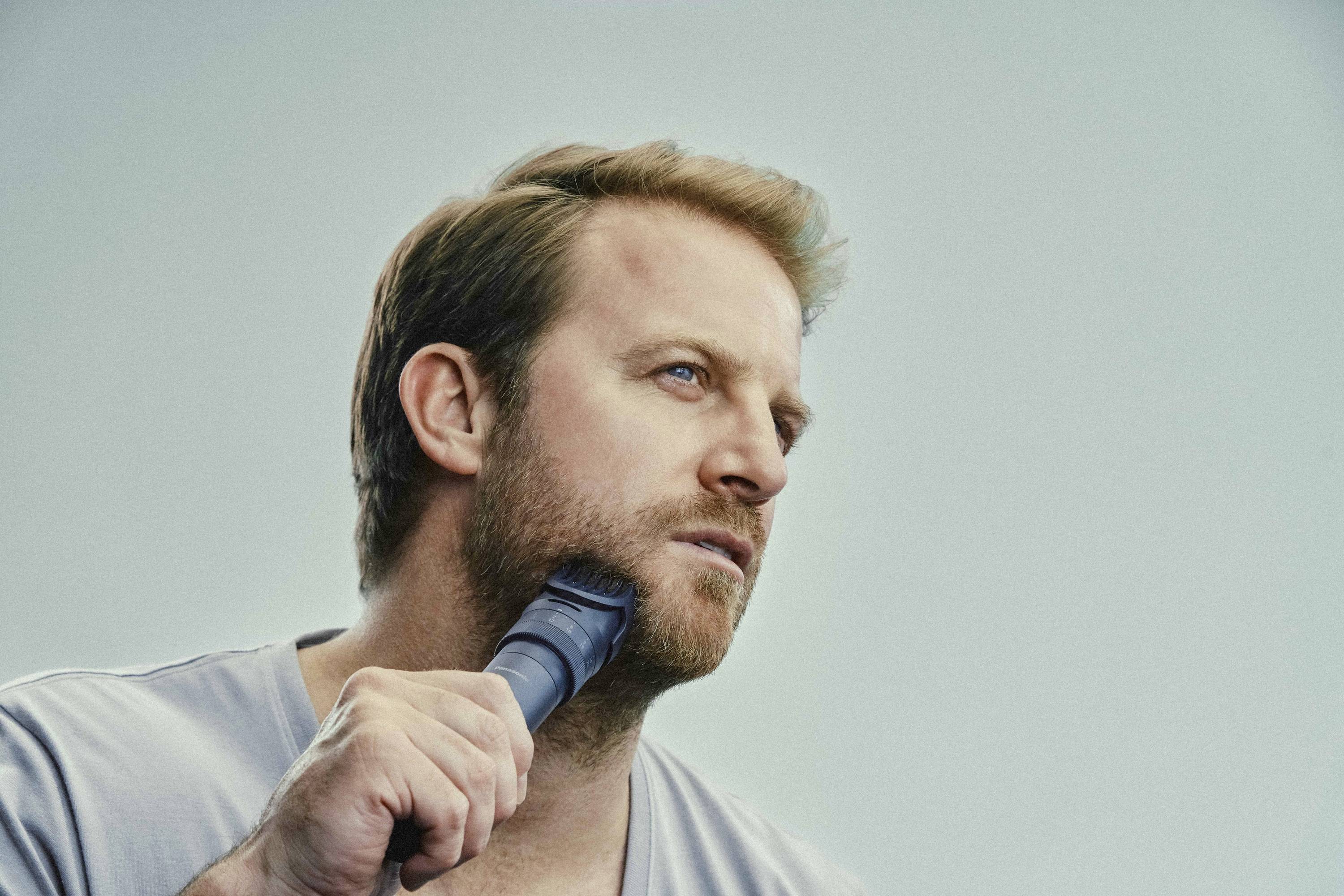 A man shaves his beard with an electric razor, his gaze thoughtfully directed to the side, against a neutral background.