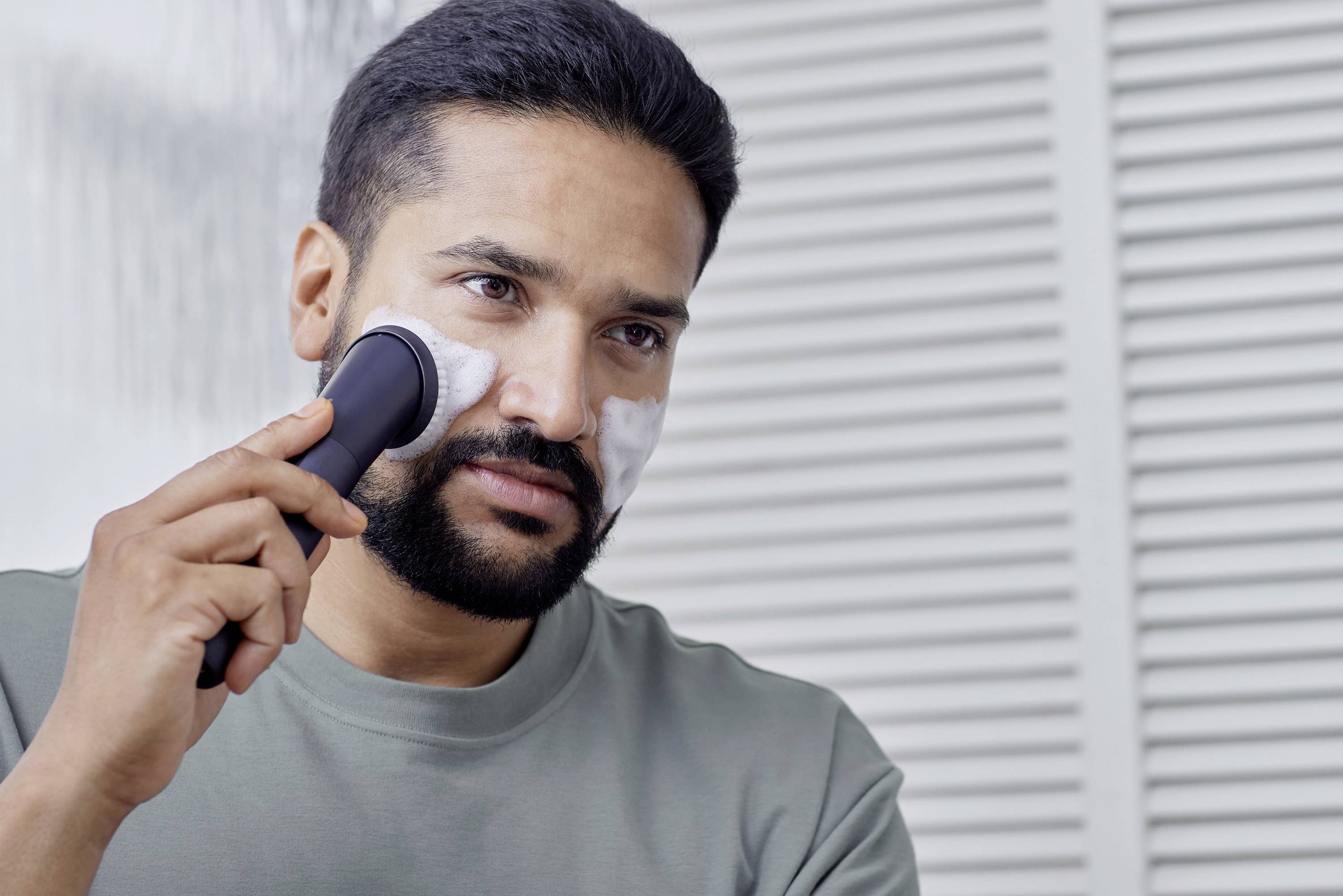 A man is shaving his face with an electric razor, with shaving foam on his cheeks. He is wearing a grey t-shirt.