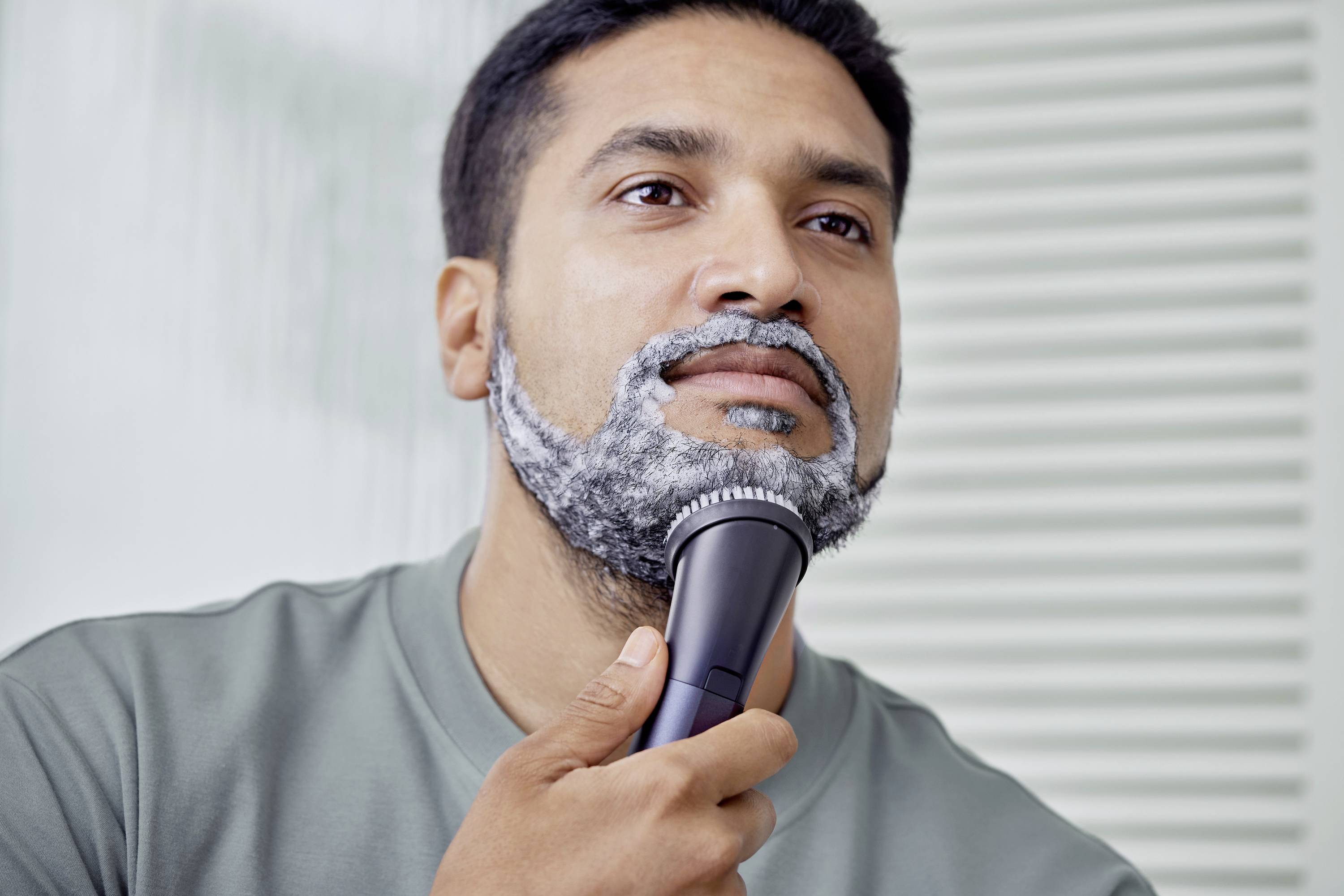 A man is shaving his beard with an electric razor. His face is covered with shaving foam.