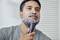 A man is shaving his beard with an electric razor. His face is covered with shaving foam.