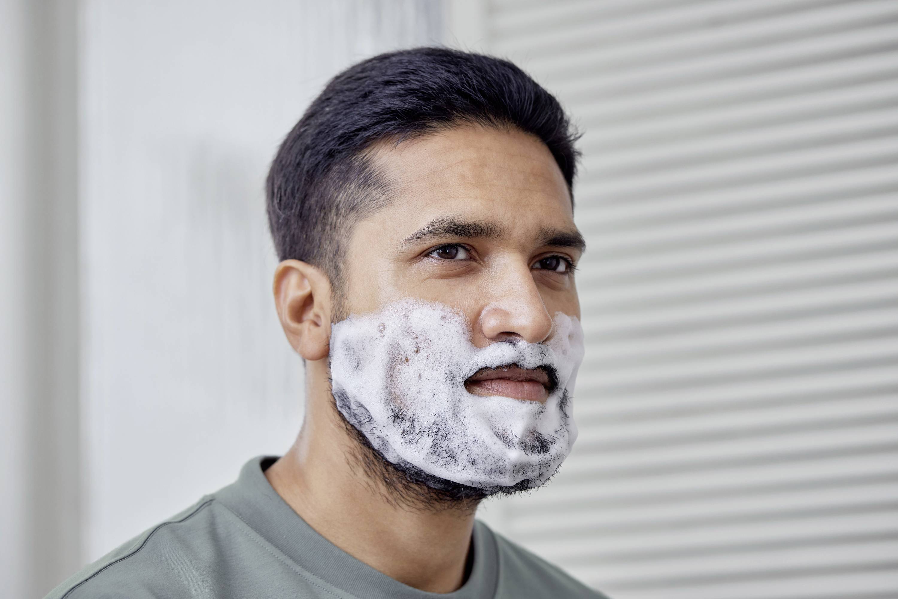 A man in a grey top has shaving foam on his face. He is standing in front of a light background with vertical blinds.