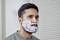A man in a grey top has shaving foam on his face. He is standing in front of a light background with vertical blinds.