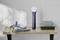 Electric facial cleansing brush with accessories and soap dish on wooden shelf, in front of white tiled wall in bathroom.