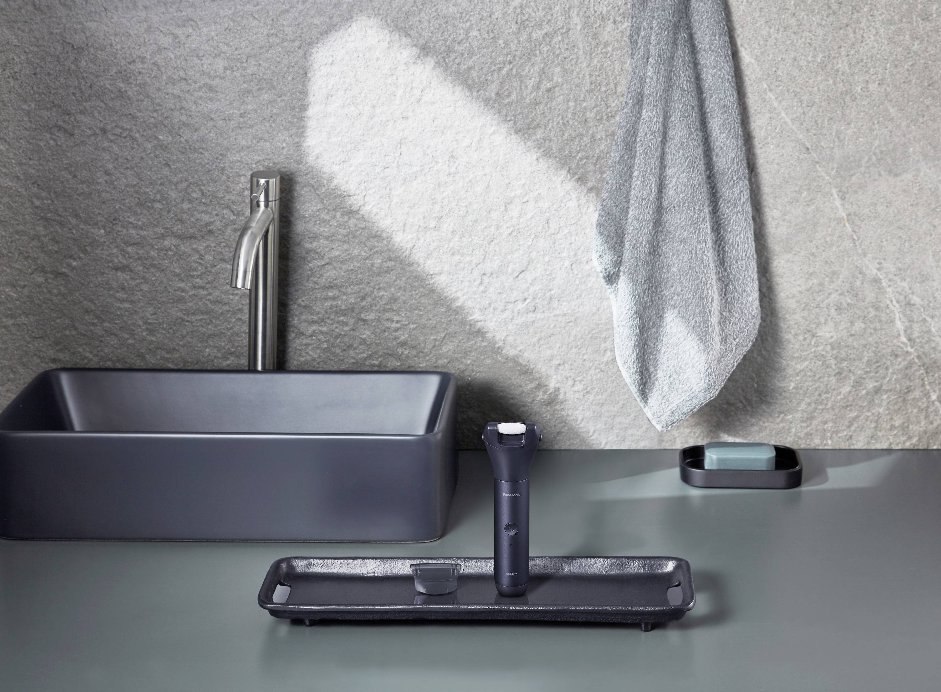 A grey bathroom with a washbasin on the left and a towel on the wall. On the table are a razor and a soap dish.