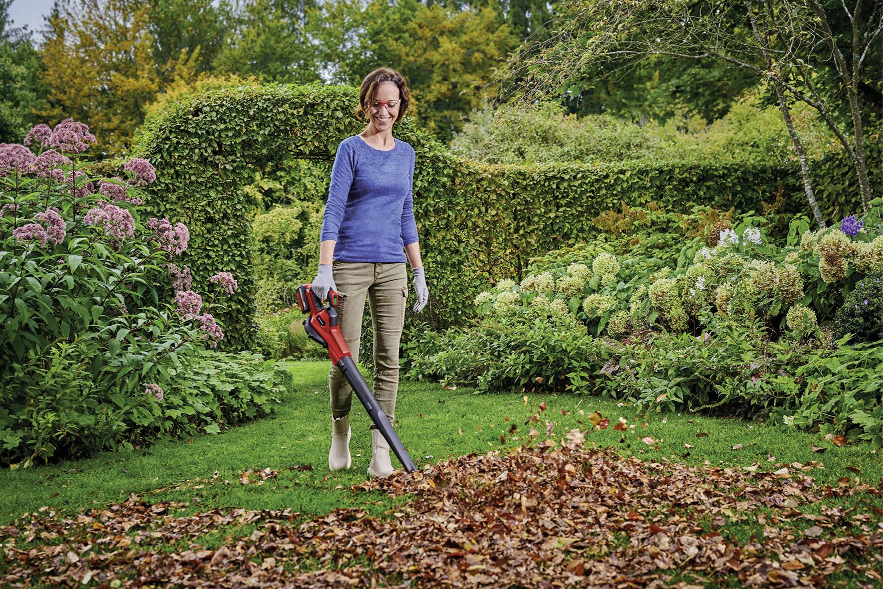A woman in gardening attire is using a leaf blower to gather a pile of autumn leaves in a lush garden area.
